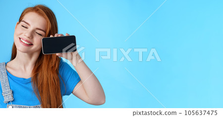 Carefree tender redhead european female tilt head, smiling closed eyes, hold smartphone horizontally, show loving girlfriend photos, romantic video online, stand blue background cheerful Carefree tender redhead european female tilt head, smiling closed eyes, hold smartphone horizontally, show loving girlfriend photos, romantic video online, stand blue background cheerful 105637475