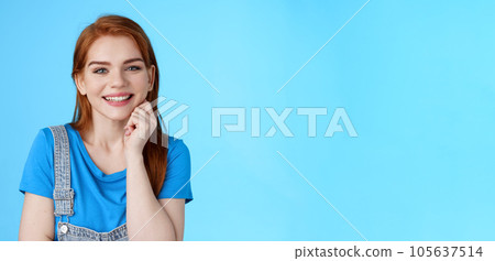 Close-up satisfied tender grinning cute redhead girl, look cute lovely camera, touch cheek smiling toothy white perfect grin, stand blue background amused, listening interesting conversation 105637514
