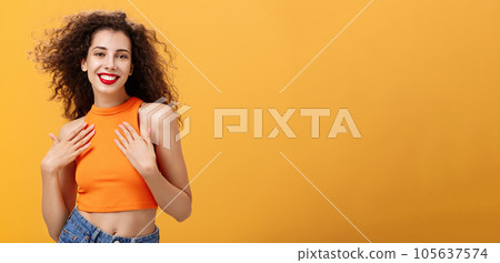 Waist-up shot of sensual and feminine tender female with curly hairstyle in cropped top holding palms on chest delighted and pleased smiling broadly posing flirty and sexy over orange wall 105637574
