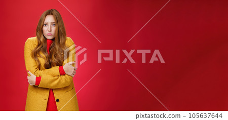 Cute and sad trembling redhead girl in light yellow coat shaking from cold, having goosebumps frowning and pouting from sorrow as freezing on windy weather over red background Cute and sad trembling redhead girl in light yellow coat shaking from cold, having goosebumps frowning and pouting from sorrow as freezing on windy weather over red background 105637644