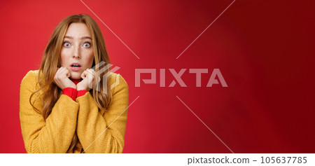 Close-up shot of shocked and scared insecure redhead woman in yellow coat pressing hands to jawline, open mouth and popping eyes at camera from amazement and fear over red background 105637785