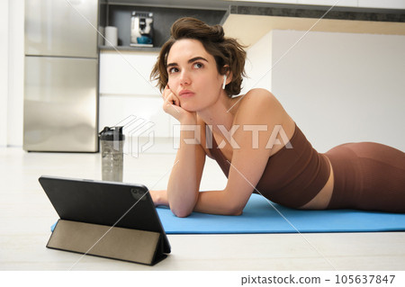 Thinking girl in sportswear, does fitness training at home, lies on rubber mat with tablet, watches workout video tutorials and looking complicated 105637847