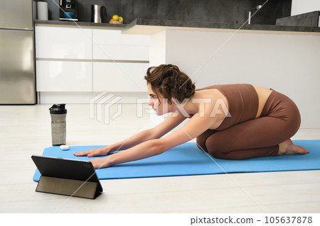 Image of young sportswoman does yoga at home, watches gym instructor tutorial video on tablet, stretches body, fitness training exercises, pilates class online from home Image of young sportswoman does yoga at home, watches gym instructor tutorial video on tablet, stretches body, fitness training exercises, pilates class online from home 105637878
