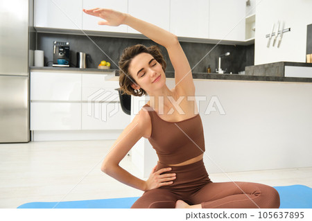 Portrait of fitness woman doing exercises in bright room at home, stretching in lotus pose, sitting on rubber yoga mat and doing workout training, drinking water from bottle Portrait of fitness woman doing exercises in bright room at home, stretching in lotus pose, sitting on rubber yoga mat and doing workout training, drinking water from bottle 105637895
