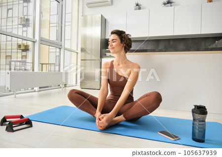 Portrait of fitness girl doing yoga on rubber mat at home, workout indoors in kitchen wearing sportswear, practice minfulness, sitting in lotus pose with water bottle next to her 105637939
