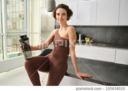 Portrait of smiling fitness girl, wearing sportswear for jogging, workout training, standing in kitchen and drinking water from sports bottle, looking fit and happy Portrait of smiling fitness girl, wearing sportswear for jogging, workout training, standing in kitchen and drinking water from sports bottle, looking fit and happy 105638010