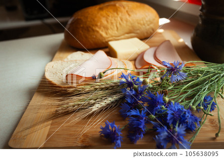 Ham sandwiches next to a loaf of bread with cereal and cornflower flowers lying on a cutting board Ham sandwiches next to a loaf of bread with cereal and cornflower flowers lying on a cutting board 105638095