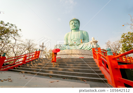 Left side view back of young Asian monk hold red umbrella and walk up on stair in front of statue 105642512
