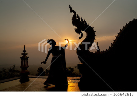 Silhouette of woman wear Thai traditional dress and stand with Thai dance in front of naga sculpture Silhouette of woman wear Thai traditional dress and stand with Thai dance in front of naga sculpture 105642613