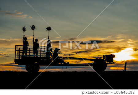Silhouette of children hold small windmill and stand on E-taen farm tractor with driver to back home Silhouette of children hold small windmill and stand on E-taen farm tractor with driver to back home 105643192