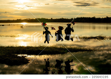 Silhouette of group of girls hold small windmill and jump together near water reservoir Silhouette of group of girls hold small windmill and jump together near water reservoir 105643195