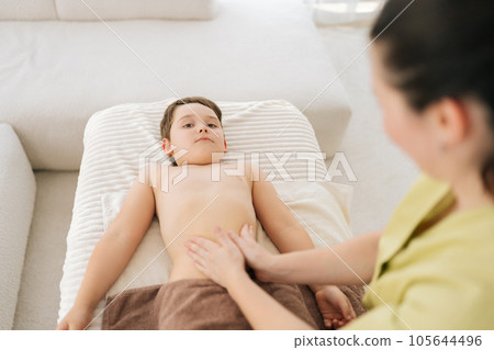 High-angle view of five year old boy patient having therapeutic massage on tummy lying on couch in medical clinic. Closeup hands of unrecognizable female pediatric masseuse massaging tummy of child. High-angle view of five year old boy patient having therapeutic massage on tummy lying on couch in medical clinic. Closeup hands of unrecognizable female pediatric masseuse massaging tummy of child. 105644496