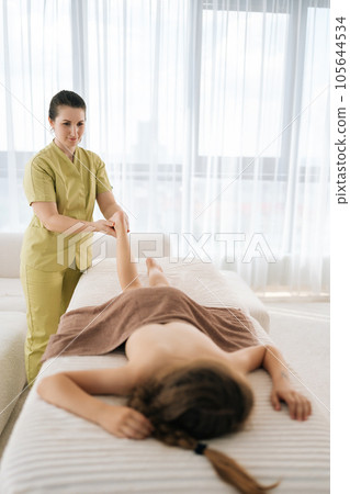 Vertical remote shot of professional female masseur doing orthopedic leg exercises of teenager girl client, prevention of pain in heel, lying on massage table in spa salon. Concept of childcare. 105644534