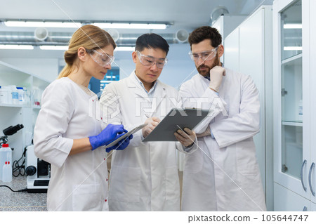 An international team of scientists and chemists standing in a laboratory in white coats and discussing work, research. They look thoughtfully at the documents with the results. An international team of scientists and chemists standing in a laboratory in white coats and discussing work, research. They look thoughtfully at the documents with the results. 105644747