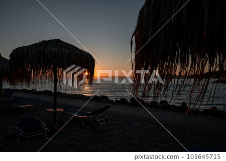 Picturesque view Sunrise From Corfu Island Overlooking Mountains Of Balkan Peninsula Of Greece, Moraitika, Corfu. In Foreground Are Silhouettes Of beach and sun loungers with umbrellas. Picturesque view Sunrise From Corfu Island Overlooking Mountains Of Balkan Peninsula Of Greece, Moraitika, Corfu. In Foreground Are Silhouettes Of beach and sun loungers with umbrellas. 105645715