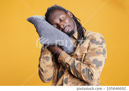 Portrait of young adult holding pillow to take a nap in studio, comfortable pose to sleep and rest. African american man with eyes closed trying to fix slpeeing schedule with bedtime hour. Portrait of young adult holding pillow to take a nap in studio, comfortable pose to sleep and rest. African american man with eyes closed trying to fix slpeeing schedule with bedtime hour. 105646692