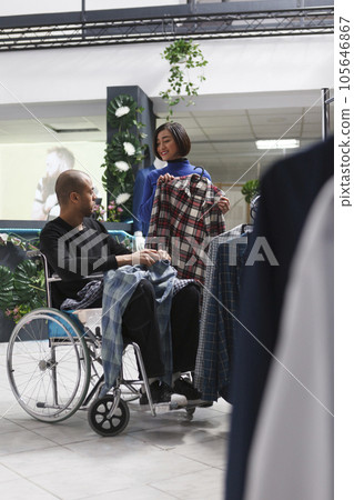 Clothing store asian woman customer showing plaid shirt on hanger to arab man with physical impairment. Mall boutique client in wheelchair getting assistance from seller while shopping for apparel Clothing store asian woman customer showing plaid shirt on hanger to arab man with physical impairment. Mall boutique client in wheelchair getting assistance from seller while shopping for apparel 105646867