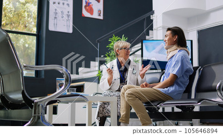 Senior medic checking patient with neck injury while asian assistant writing medical expertise on clipboard during checkup visit in hospital lobby. Injured man wearing cervical collar in waiting area Senior medic checking patient with neck injury while asian assistant writing medical expertise on clipboard during checkup visit in hospital lobby. Injured man wearing cervical collar in waiting area 105646894
