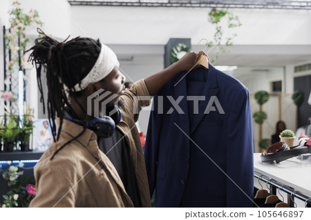 African american man holding blazer in clothing store, examining fabric and size. Customer looking at formal jacket on hanger while choosing new outfit in shopping center boutique 105646897