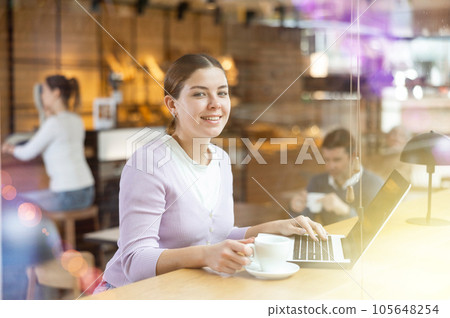 Portrait of focused young female freelancer surfing Internet on notebook while drinking tea or coffee in cafeteria 105648254