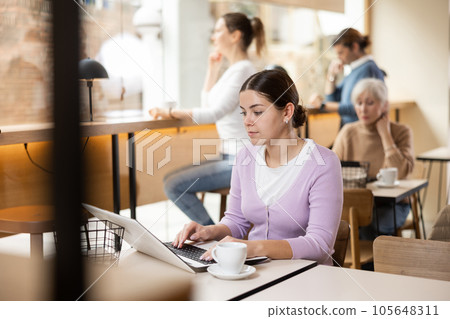 Positive European young woman drinking and enjoying coffee while working on laptop in cafe. Freelance and remote work Positive European young woman drinking and enjoying coffee while working on laptop in cafe. Freelance and remote work 105648311