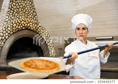 Latino woman taking out baking tray with pizza from oven in kitchen of pizzeria 105648377