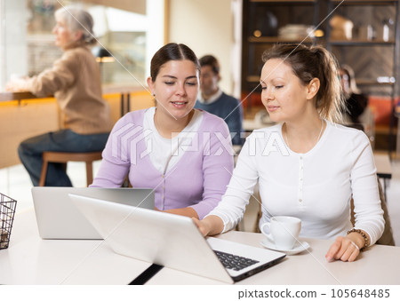 Two young women having fun together looking at laptop screen while sitting at the bar of restaurant 105648485