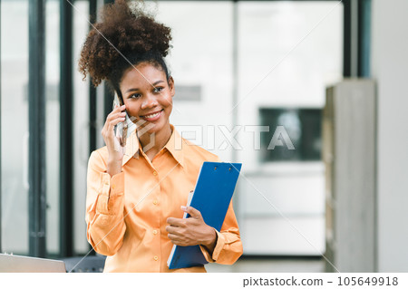 Portrait of a young, smiling, and cheerful African American businesswoman working in a casual office, holding a financial report folder while making a business call. Portrait of a young, smiling, and cheerful African American businesswoman working in a casual office, holding a financial report folder while making a business call. 105649918
