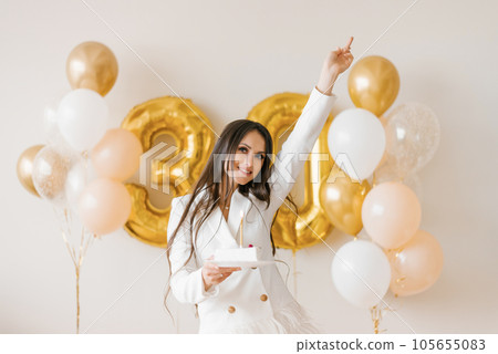 Young caucasian woman holding a cake with a candle in honor of the thirtieth birthday in a stylish white dress with feathers 105655083