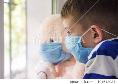 Sad child and his teddy bear both in protective medical masks looks out window.  105658081