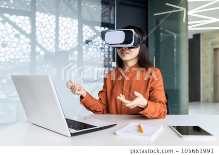 Asian woman wearing virtual reality glasses sitting at a desk in a workspace inside the office, businesswoman in online meeting talking remotely with colleagues, virtual meeting simulation. 105661991