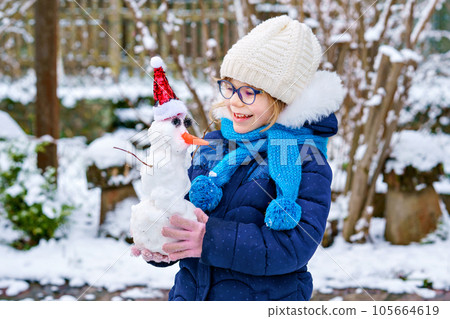 Cute little preschool girl with glasses making mini snowman. Adorable healthy happy child playing and having fun with snow, outdoors on cold day. Active leisure with children in winter 105664619