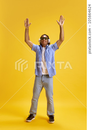 Full-length image of young cheerful man in sunglasses and casual clothes listening to music in headphones against yellow studio background 105664624