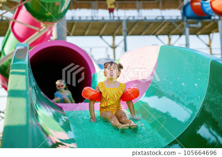 Little preschool girl on water slide in aqua park. Happy child having fun on water slides on family summer vacation in tropical resort. Amusement park with wet playground for young child and baby. Little preschool girl on water slide in aqua park. Happy child having fun on water slides on family summer vacation in tropical resort. Amusement park with wet playground for young child and baby. 105664696