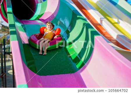 Little preschool girl on water slide in aqua park. Happy child having fun on water slides on family summer vacation in tropical resort. Amusement park with wet playground for young child and baby. Little preschool girl on water slide in aqua park. Happy child having fun on water slides on family summer vacation in tropical resort. Amusement park with wet playground for young child and baby. 105664702