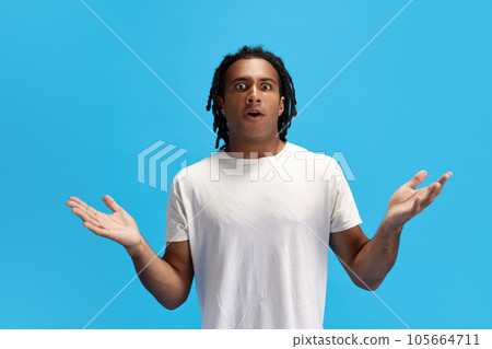 Young african man with dread spreading hands and standing with shocked, surprised face against blue studio background 105664711