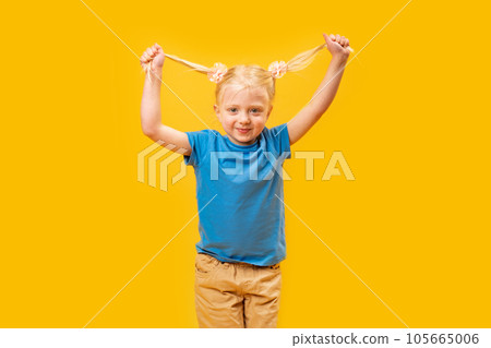Portrait of funny little girl in blue t-shirt having fun and holding two ponytails with her blonde hair. Yellow background Portrait of funny little girl in blue t-shirt having fun and holding two ponytails with her blonde hair. Yellow background 105665006
