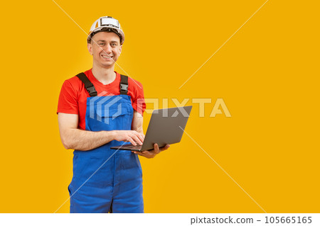 Young man in safety helmet and blue overalls holds laptop in hands looks at the camera and smiles. Isolated on yellow background. Copy space. Young man in safety helmet and blue overalls holds laptop in hands looks at the camera and smiles. Isolated on yellow background. Copy space. 105665165