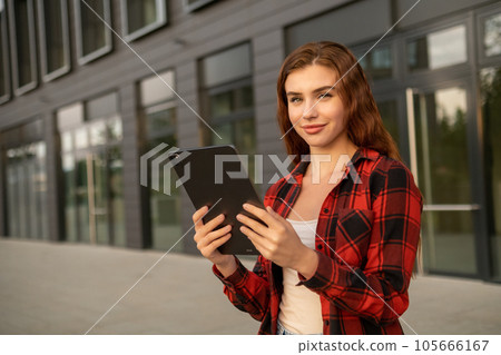 Portrait of the young pretty red haired woman holding a tablet standing near modern building.  105666167