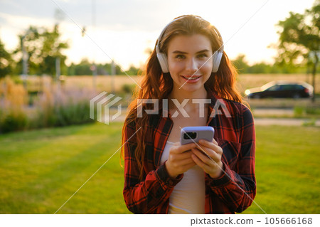Happy young woman wearing wireless headphones for listening to the music via mobile phone in the park at sunset. 105666168