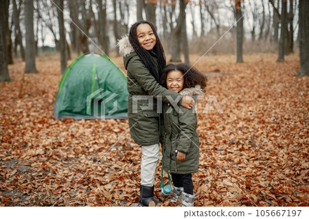 Beautiful little black girls standing near tent in the forest. Two little sisters relaxing in autumn forest. Black girls wearing khaki coats. 105667197