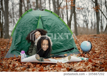 Beautiful little black girls in tent camping in the forest. Two little sisters lying in a tent in autumn forest and looking on a map. Black girls wearing khaki coats. 105667202