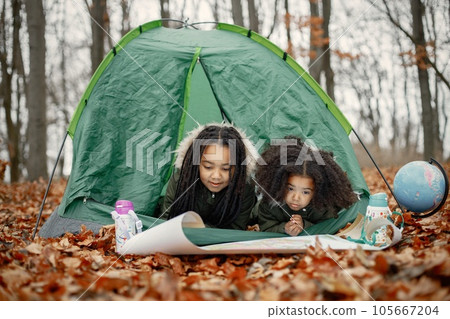 Beautiful little black girls in tent camping in the forest. Two little sisters lying in a tent in autumn forest and looking on a map. Black girls wearing khaki coats. 105667204