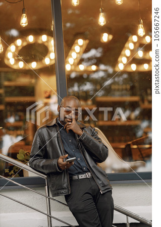 Black man standing on a street in a city. Man wearing sunglasses and a leather jacket. Business man using a phone. Black man standing on a street in a city. Man wearing sunglasses and a leather jacket. Business man using a phone. 105667246