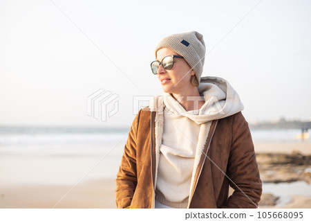 Portrait of young stylish woman wearing brown padded jacket, hoodie, wool cap and sunglasses on long sandy beach in spring 105668095