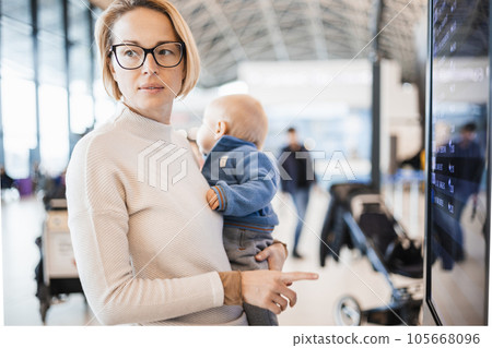 Mother traveling with child, holding his infant baby boy at airport terminal, checking flight schedule, waiting to board a plane. Travel with kids concept 105668096