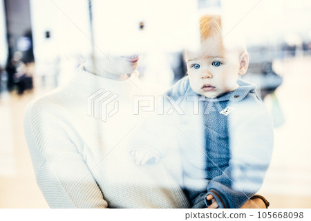 Thoughtful young mother looking trough window holding his infant baby boy child while waiting to board an airplane at airport terminal departure gates. Travel with baby concept Thoughtful young mother looking trough window holding his infant baby boy child while waiting to board an airplane at airport terminal departure gates. Travel with baby concept 105668098