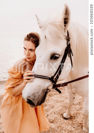 A woman in a dress stands next to a white horse on a beach, with the blue sky and sea in the background. A woman in a dress stands next to a white horse on a beach, with the blue sky and sea in the background. 105668589