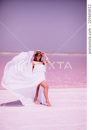 Woman in pink salt lake. She in a white dress and hat enjoys the scenic view of a pink salt lake as she walks along the white, salty shore, creating a lasting memory. 105668651