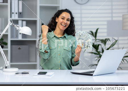 A young Latin American woman is sitting at the desk in the office and holding cash money in her hands. Rejoices and smiles at the camera, shows a victory hand gesture yes. A young Latin American woman is sitting at the desk in the office and holding cash money in her hands. Rejoices and smiles at the camera, shows a victory hand gesture yes. 105673221